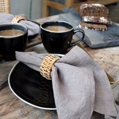 A set of taupe linen napkins displayed on a wooden table alongside black cups with coffee, indicating a sophisticated dining setting.