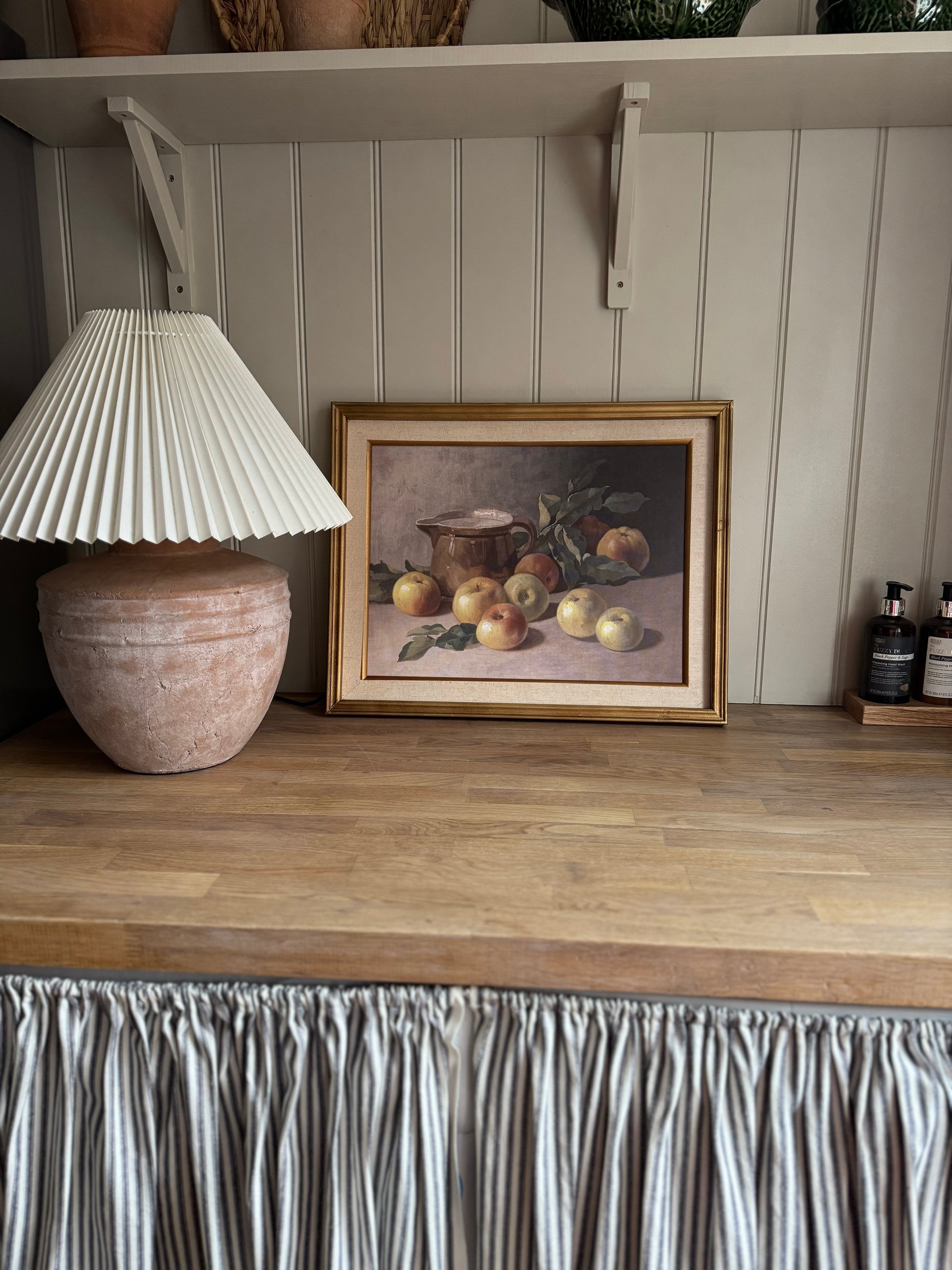 Framed still life painting of apples on a wooden shelf with a lamp and decorative items.