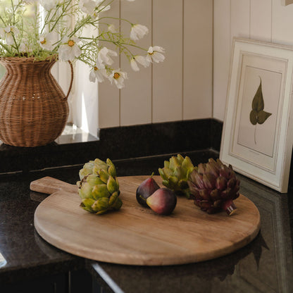 Wooden cutting board with fruits and a vase of flowers on a kitchen counter.