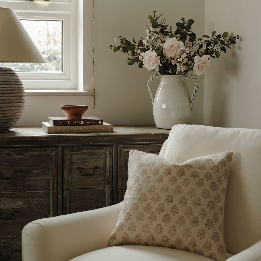 Cozy living room with a white armchair, floral arrangement in a vase, and books on a wooden cabinet.