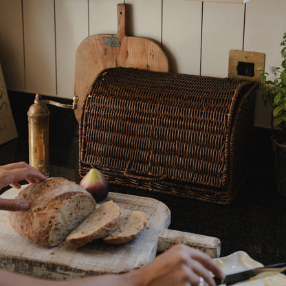 Person slicing bread on a wooden cutting board with a wicker bread box in the background.
