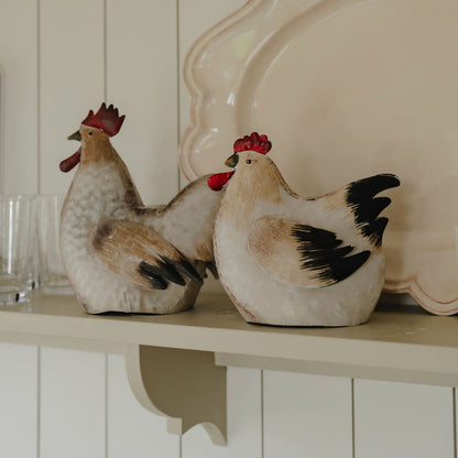 Two ceramic chicken figurines on a shelf with a decorative plate in the background.