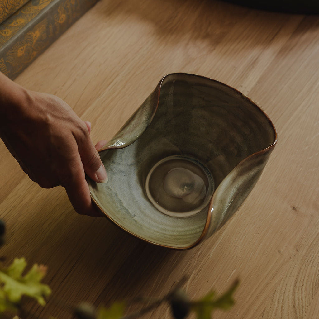 A hand holds a handmade ceramic bowl with a wavy rim and a textured, earthy green-brown glaze, set on a light wooden table with books and a tray in the background.