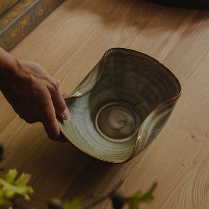 A hand holds a handmade ceramic bowl with a wavy rim and a textured, earthy green-brown glaze, set on a light wooden table with books and a tray in the background.