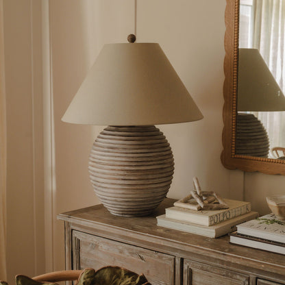 A Saturn Table Lamp with a modern geometric base and a beige linen shade, placed on a wooden side table in a room.