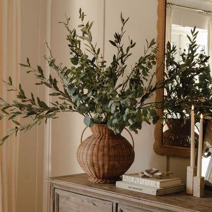 A woven wicker vase with a plant arrangement on a wooden surface, possibly a dresser or table, in a room with a mirror and chair.