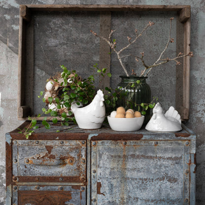 Decorative setting with ceramic chickens, a bowl of eggs, and greenery on a rustic metal surface.